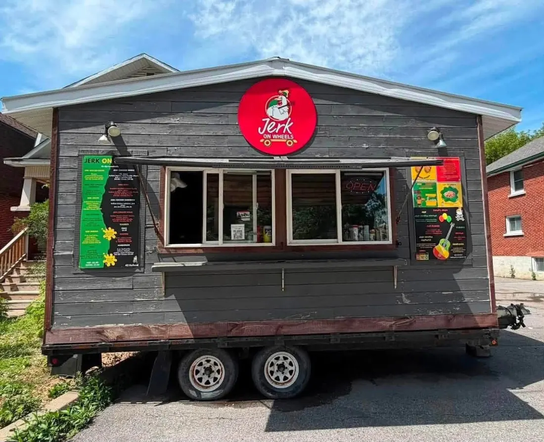 Jerk on Wheels food truck with grey wood cladding, a round red logo and a serving window, parked on a sunny street