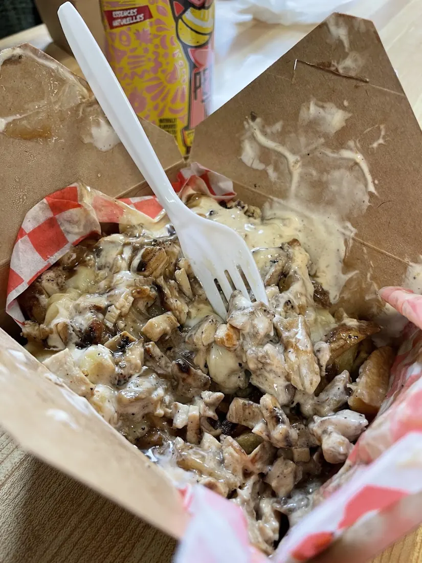 Jerk chicken poutine in a takeout box with melted cheese, fries and a plastic fork