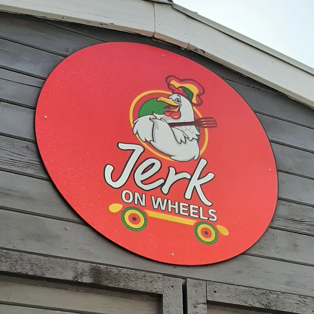 Round red Jerk on Wheels logo with a cartoon rooster on a skateboard, on grey wood